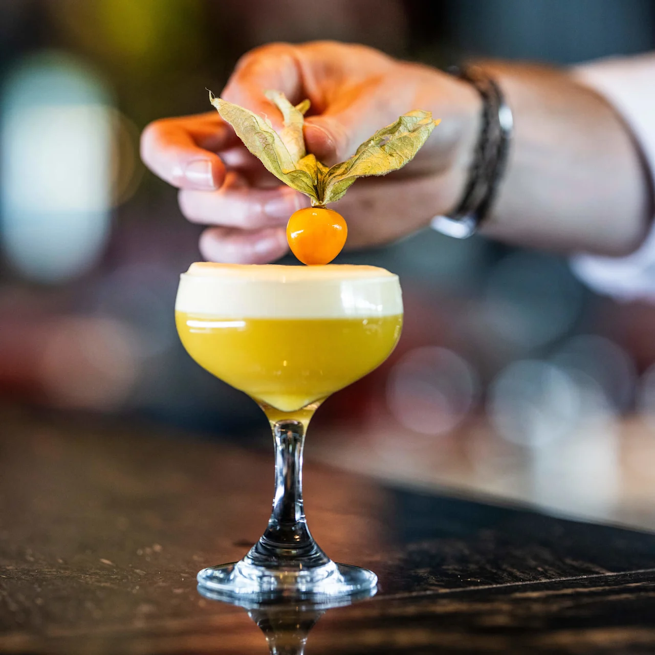 Ett cocktailglas med en skummig gul drink garneras med en physalis av en hand i en suddig barmiljö.