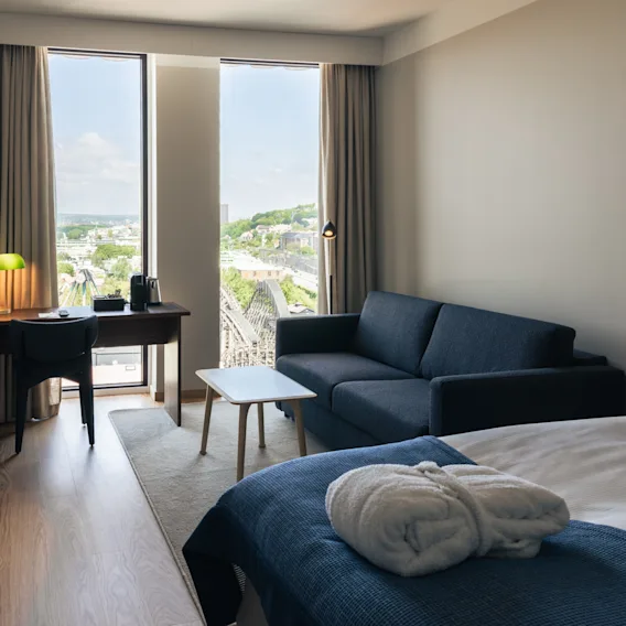 Ett ljust och inbjudande hotellrum med en mysig säng, en soffa, ett skrivbord och stora fönster med stadutsikt.