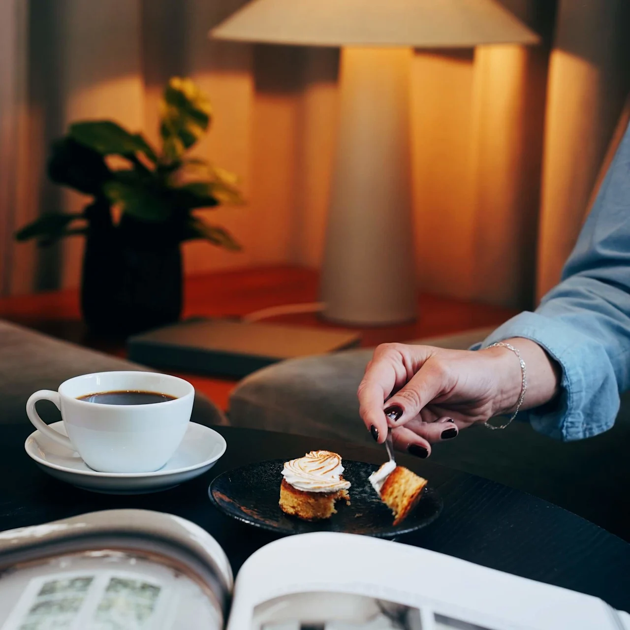 En person skär en bit tårta på en tallrik bredvid en kaffekopp, med en tänd lampa och planta i bakgrunden.