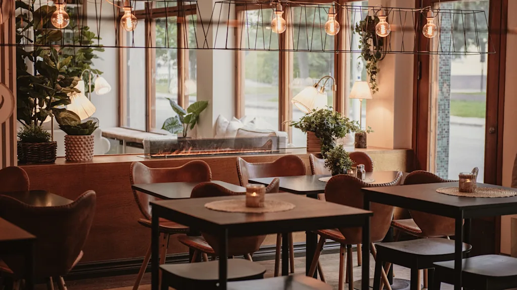 A warm and inviting restaurant interior with tables, chairs, plants, and soft lighting, creating a cozy atmosphere.