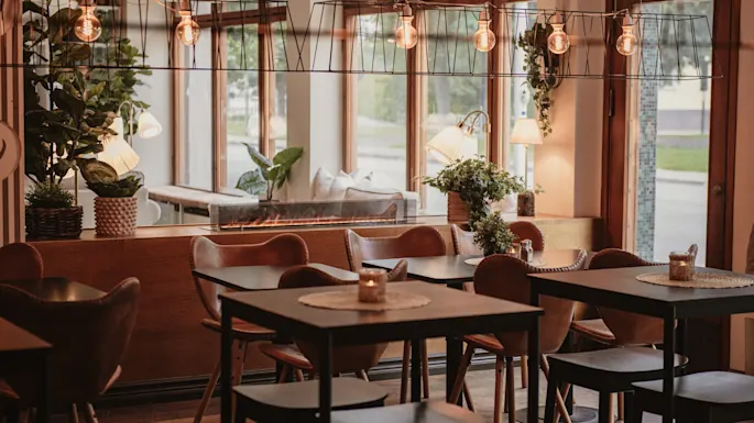 Et varmt og innbydende restaurantinteriør med bord, stoler, planter og myk belysning, som skaper en koselig atmosfære.