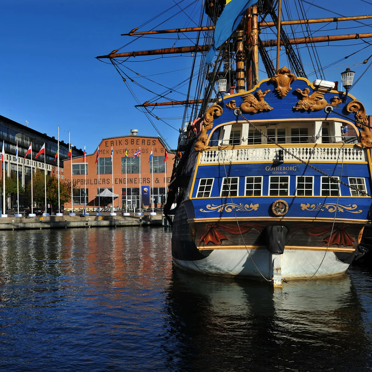 A large blue and gold ship named Götheborg docks beside a waterfront lined with flags and modern buildings. Nearby, "Eriksbergs Mek. Verkstads AB" is visible on a brick structure.