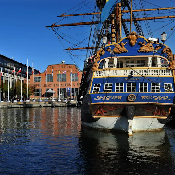 Ett stort blått och guld skepp vid namn Götheborg är förtöjt vid en kaj kantad av flaggor och moderna byggnader. I närheten syns "Eriksbergs Mek. Verkstads AB" på en tegelbyggnad.