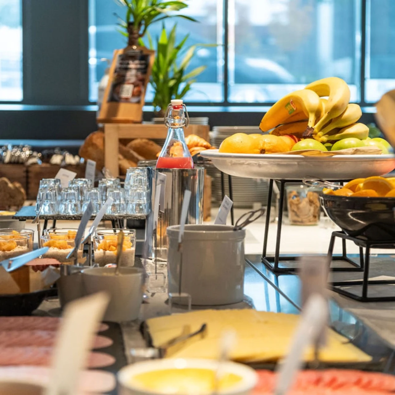 Assorted breakfast buffet with sliced meats, cheeses, fruits, and beverages arranged on a long table in a well-lit dining area with large windows.