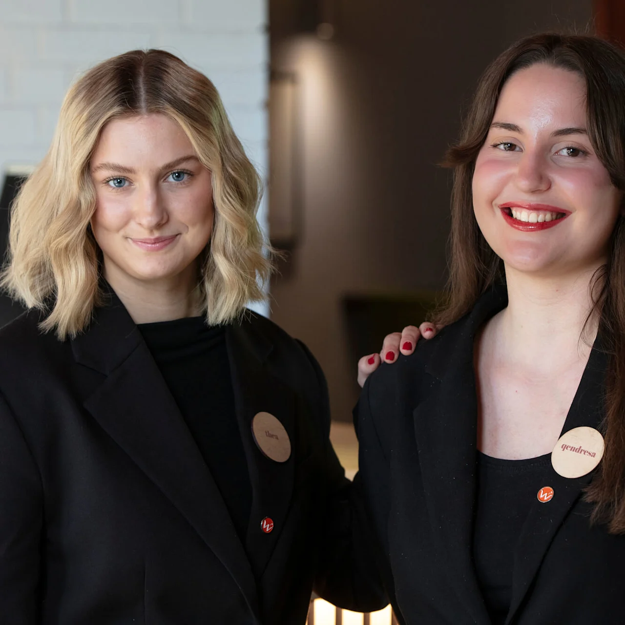 Two smiling women in black blazers, looking at the camera, indoors with a white wall behind them.