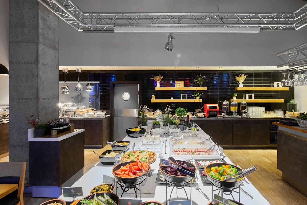 Buffet table displaying assorted salads, fruits, and pastries; situated in a modern dining area with wooden floors and decorative shelves, under bright lighting.