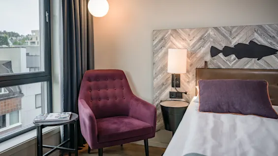 Et moderne hotellrom med en komfortabel seng, en lilla lenestol ved vinduet, og en dekorativ fiskesilhuett på sengegavlen.