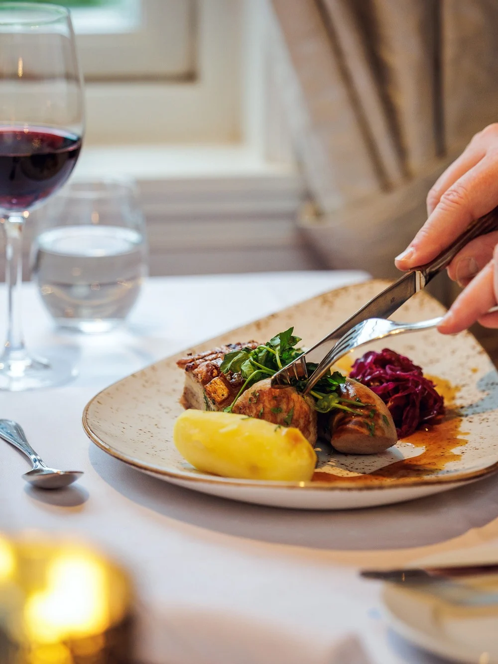 A person cuts into a meal on a white plate, surrounded by a glass of red wine and a lit candle, at a table beside a window.