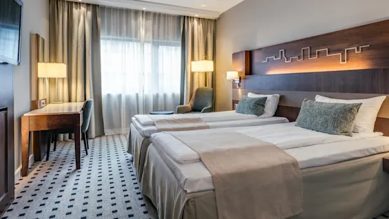Twin beds with gray pillows and beige throws, positioned in a modern hotel room. There’s a desk with a chair and lamp beside a large window with curtains.