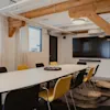 A modern meeting room with a long white table, black and yellow chairs, a large TV screen, and exposed wooden beams. Text: MADAM.