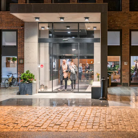 En person lämnar en modern ingång i glas och pratar med en annan i vinterkappa; inne konverserar flera personer i ett varmt belyst rum. Våta kullerstenar reflekterar ljuset utanför.