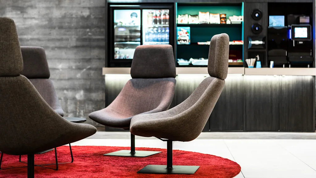 Grå lounge-stole står på et rødt tæppe foran en snackbar med et køleskab og hylder med snacks og drikkevarer. Tekst: "Lounge 24".