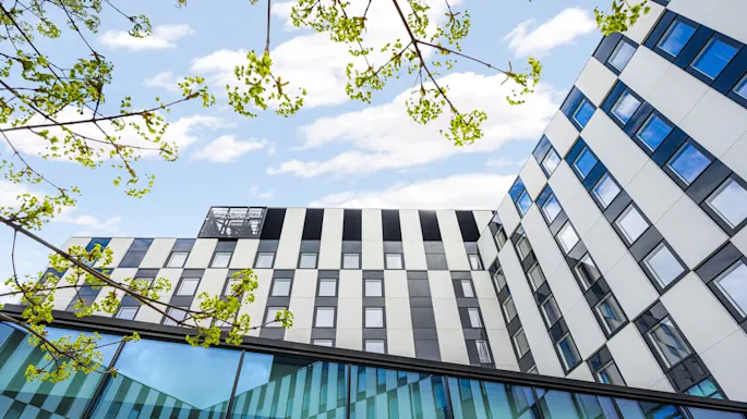 Byggnad med geometriska mönster står under en blå himmel. Gröna grenar ramar in utsikten och skapar kontrast med arkitekturen. Överst står "CLARION HOTEL & CONGRESS."