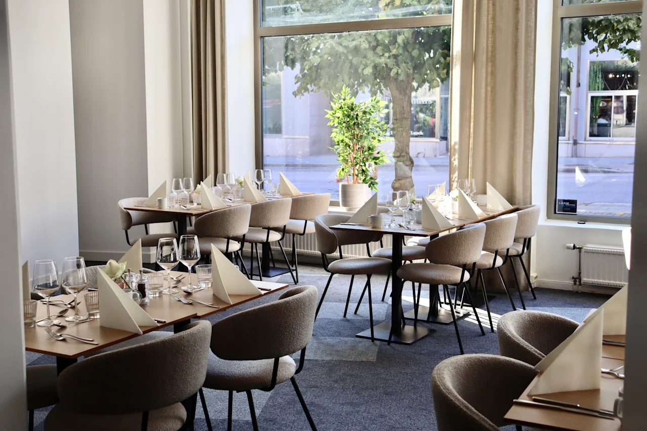 Tables are elegantly set with glasses and folded napkins in a modern restaurant. Soft chairs surround the tables, and large windows offer a view of trees outside.