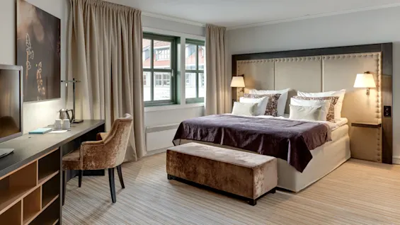 En dubbelsäng med mjuka kuddar står i ett modernt hotellrum, flankerad av lampor, med ett skrivbord och en stol i närheten, beige gardiner och konstverk på väggarna.