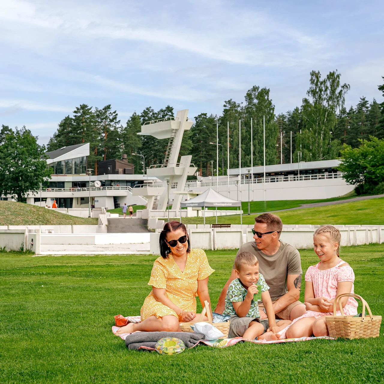 A family sits on a picnic blanket, eating and engaging in conversation. They are surrounded by a sprawling green lawn with trees and modern buildings in the background.