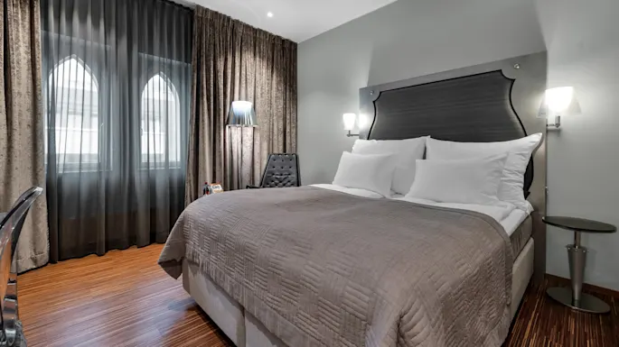 A neatly made double bed with gray and white bedding, placed beside a small nightstand and chair, is illuminated by soft wall lights in a modern, well-furnished bedroom.