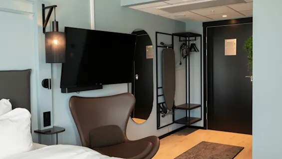 Et moderne hotellrom med seng, lenestol, veggmontert TV, speil og et klesstativ med strykebrett.