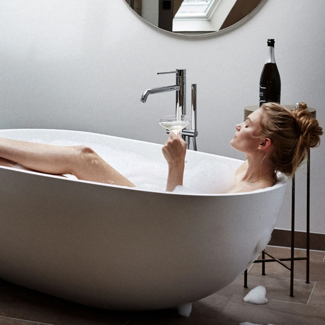 Woman relaxing in a bubble bath, holding a wine glass, in a modern bathroom with a round mirror and champagne bottle on a stand.