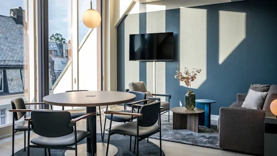 Runt träbord omgivet av fyra stolar, upplyst av hängande lampa; beläget nära stora fönster med takutsikt och intill en loungedel med soffa, TV och dekorativt bord.