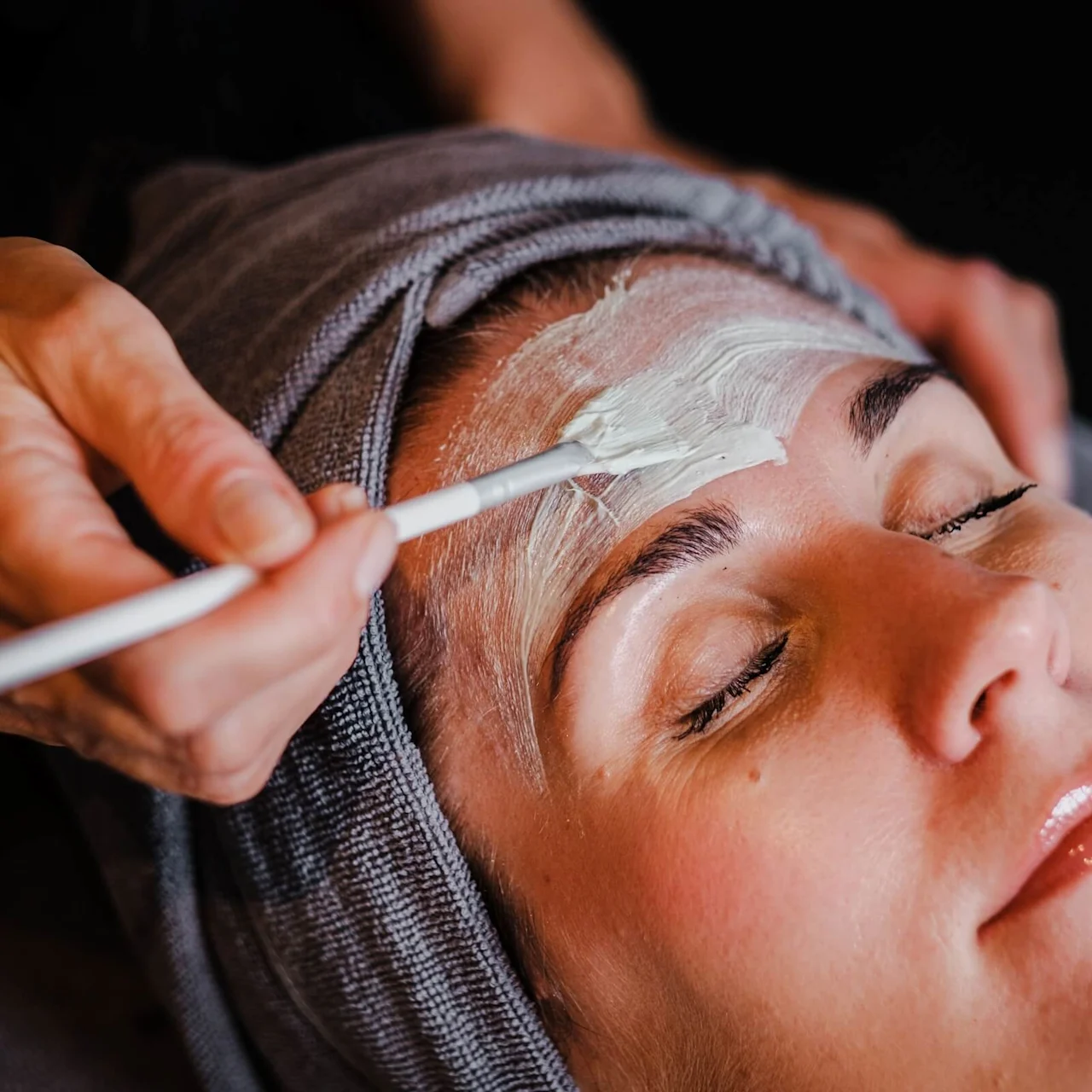 A person relaxes as cream is applied to their forehead with a brush by another person. The setting is a spa-like environment, with a towel wrapped around the head.