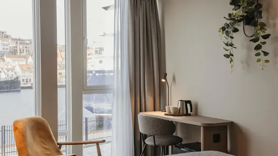 Et hyggeligt hotelværelse med skrivebord, lænestol og et stort vindue med naturskøn udsigt over en by ved vandet.