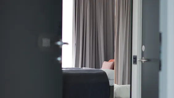 En utsikt inn i et hotellrom med en seng og gardiner, sett gjennom en delvis åpen dør.