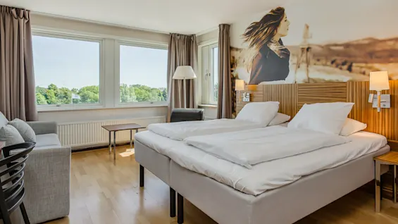 Siististi pedattu parisänky valkoisilla liinavaatteilla sijaitsee hotellihuoneessa, jossa on suuret ikkunat, vihreä näkymä, puulattia, seinämaalaus ja yöpöydän lamput.