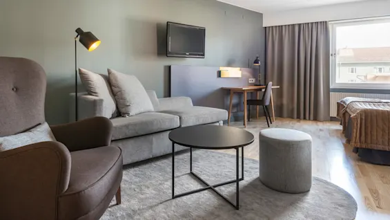 Ett vardagsrum har moderna möbler, inklusive en grå soffa, fåtölj och runt soffbord, på en matta. Ett skrivbord och TV finns i bakgrunden med ett stort fönster.