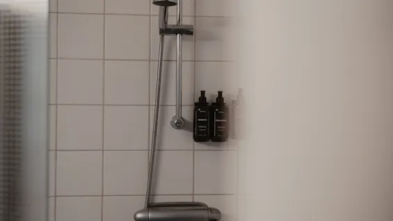 A modern shower with white tiled walls, featuring a shower head, control panel, and wall-mounted shampoo and conditioner bottles at Home Hotel Uppsala.