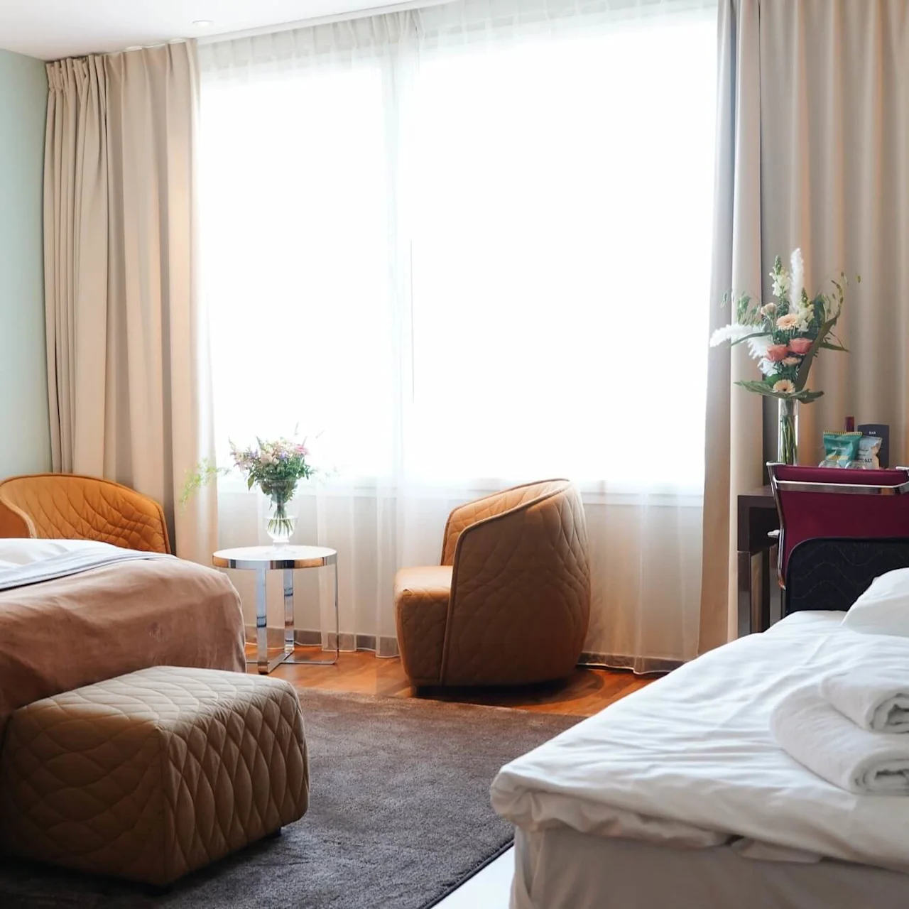 A cozy hotel room features two large beds draped with brown blankets. Orange chairs and a glass table with flowers sit near a bright window, creating a welcoming atmosphere.