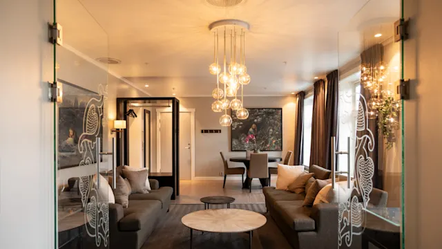 Lystekroner lyser opp en moderne stue med to sofaer, et rundt salongbord og dekorative glassdører; en spisestue med kunst på veggene sees i bakgrunnen.