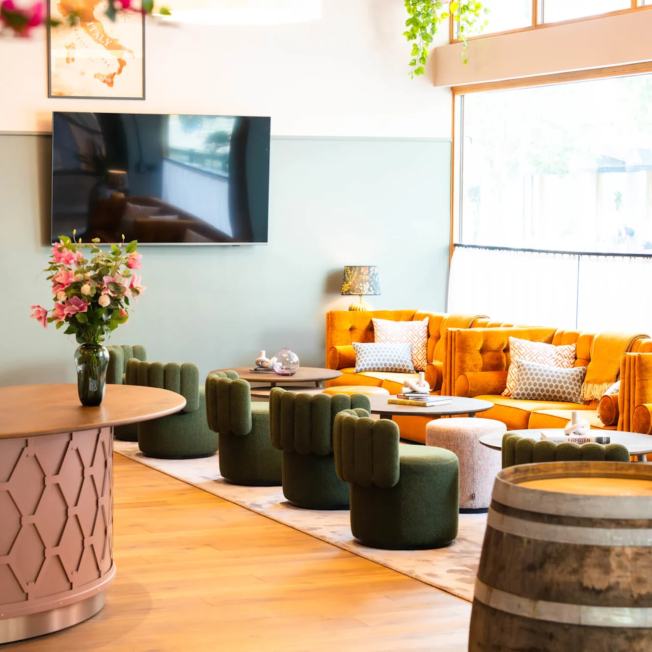 A reception desk with a vase of pink flowers is in front of green chairs and mustard sofas. A wall-mounted TV, paintings, and large windows with natural light complete the cozy lounge setting.