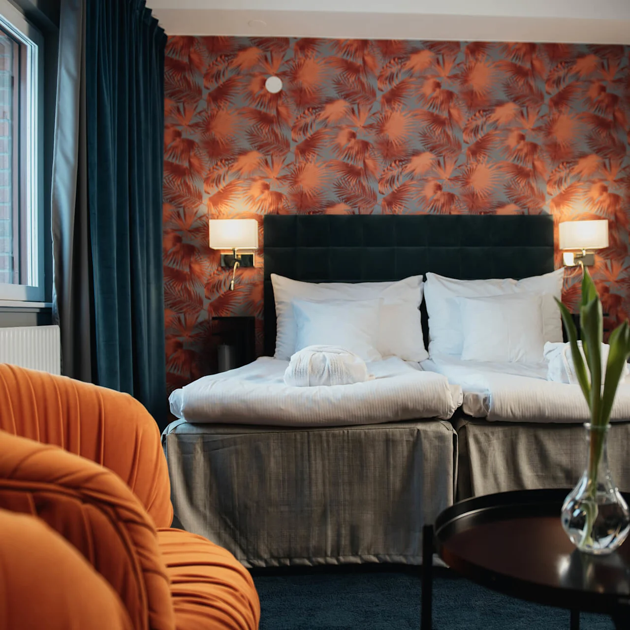 A bed with white linens and towels sits against a red, leafy patterned wall; nearby, an orange chair and small table with a vase add accents to the cozy room.
