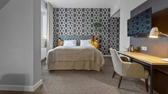 Ett modernt hotellrum med en bekväm säng, en stilfull mönstrad accentvägg och en funktionell skrivbordsplats med TV.
