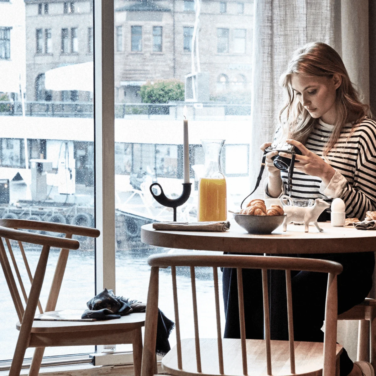 Woman photographing breakfast table with croissants, juice, and candle by a window. Boats and buildings visible outside in a cozy café setting.