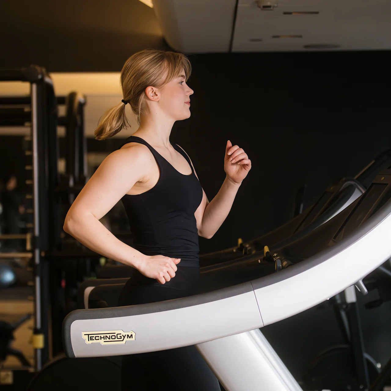 A person is jogging on a treadmill in a gym, wearing workout attire. The treadmill shows the brand "TechnoGym." Gym equipment and mirrors are visible in the background.