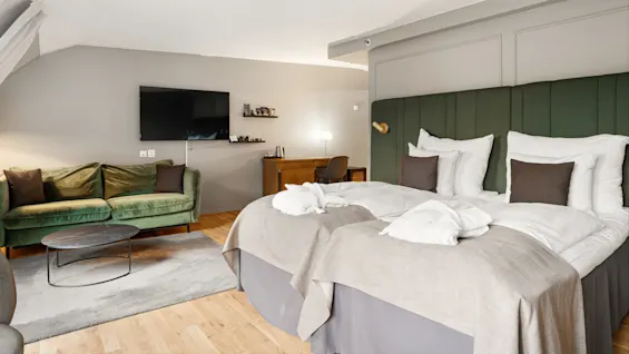To pænt redte enkeltsenge står i et moderne hotelværelse med en grøn sofa, et fladskærms-tv på væggen, et lille træskrivebord og et rundt sofabord.