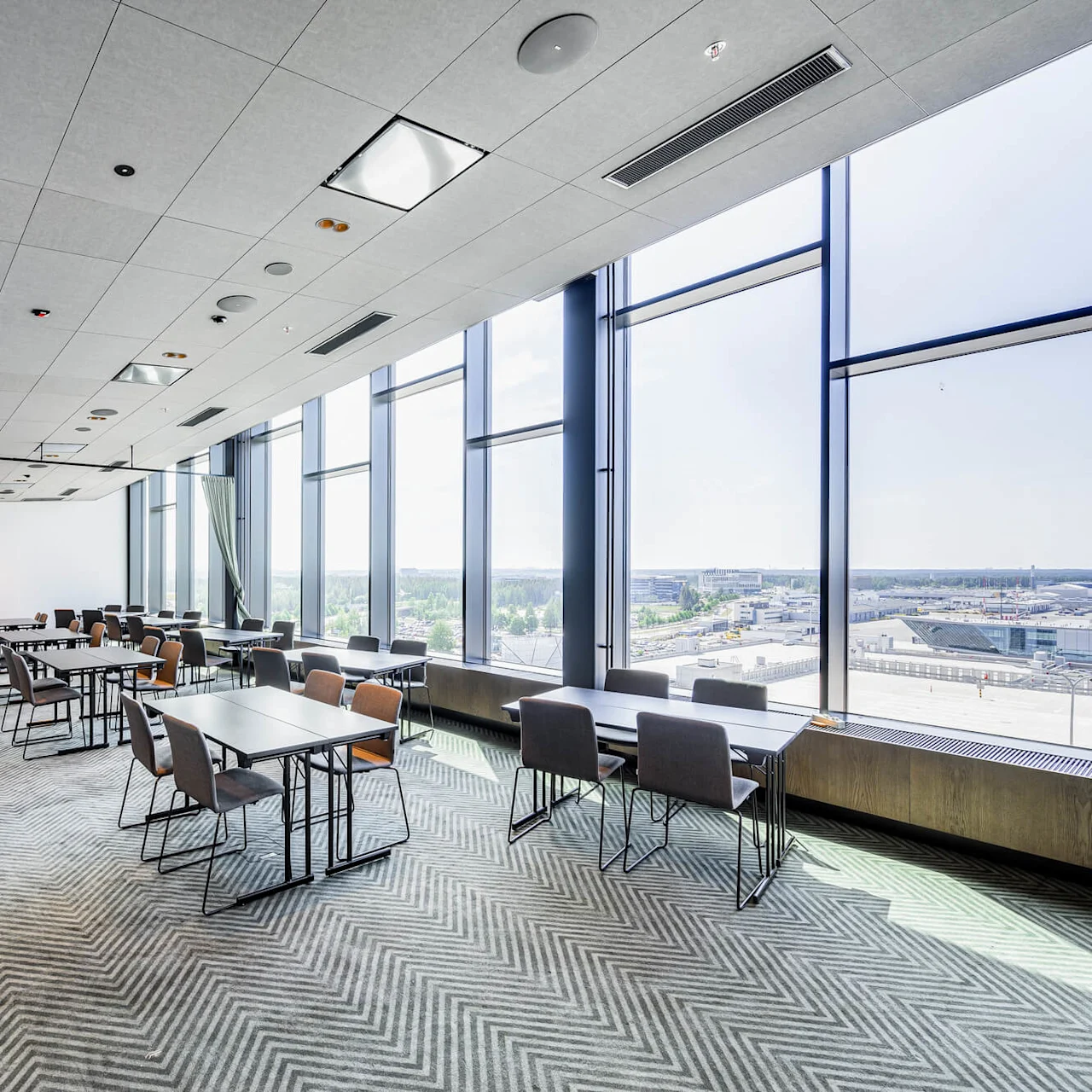 Tables and chairs are arranged in a spacious, bright conference room with large windows overlooking a cityscape. The room features light-colored walls and ceiling with patterned carpeting.