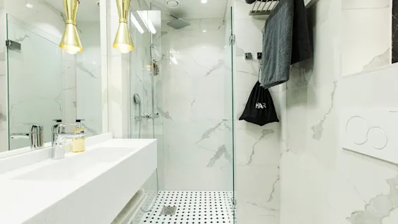 A modern bathroom features a white marble shower with glass door, black-and-white tile floor, two hanging gold lights above a counter with soap, and a hanging black bag labeled "HÅR".