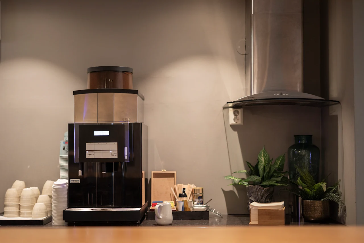 En kaffemaskin står på en benk, omgitt av engangskrus og potteplanter. I nærheten er en avtrekkshette festet til veggen. Omgivelsene synes å være en kafé.