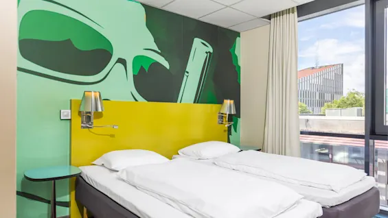 Two white twin beds stand beside a yellow headboard with attached lamps, against a vibrant wall mural depicting sunglasses, in a bright, modern room with a large window.