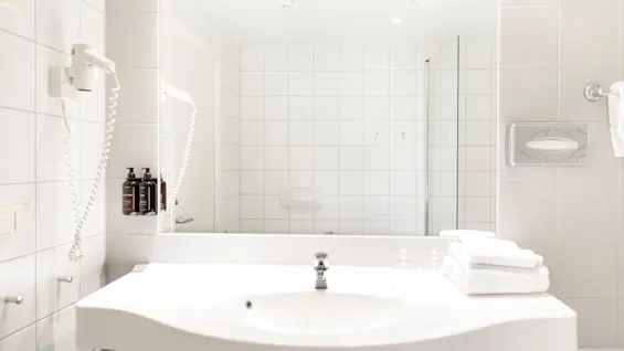 Et rent og moderne hotellbad med hvit vask, stort speil, hårføner og friske håndklær, klart til bruk.