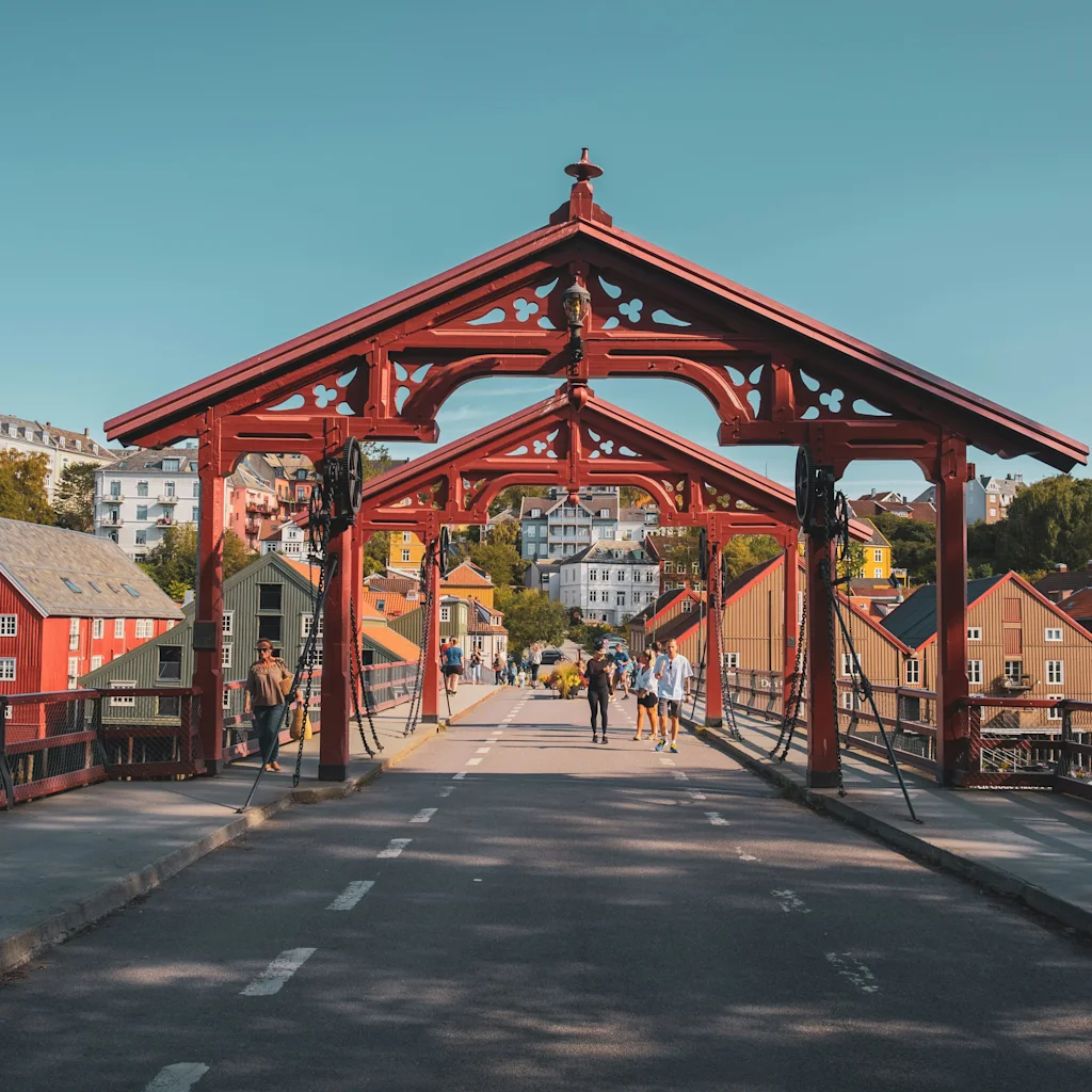 Ihmiset kävelevät punaisen puusillan yli kohti kaupunkia, jossa on värikkäitä rakennuksia kirkkaan sinisen taivaan alla.