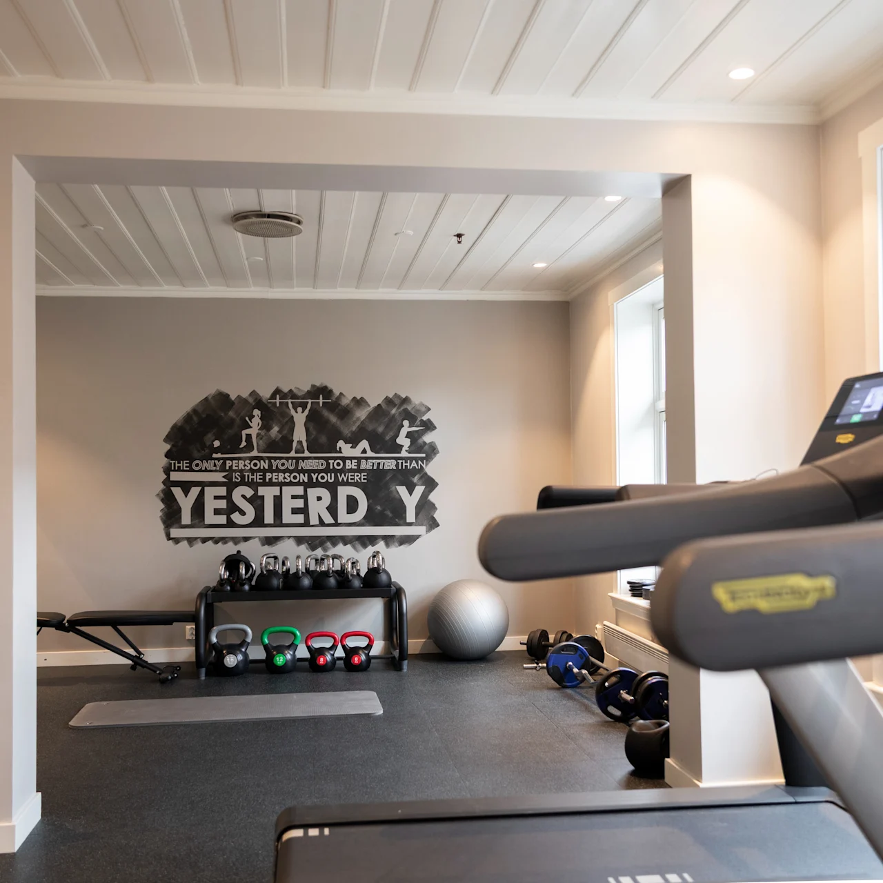 Ett löpband riktas mot flera gymutrustningar som kettlebells och en balansboll i ett ljust träningsrum. Text på väggen lyder: "THE ONLY PERSON YOU NEED TO BE BETTER THAN IS THE PERSON YOU WERE YESTERDAY."