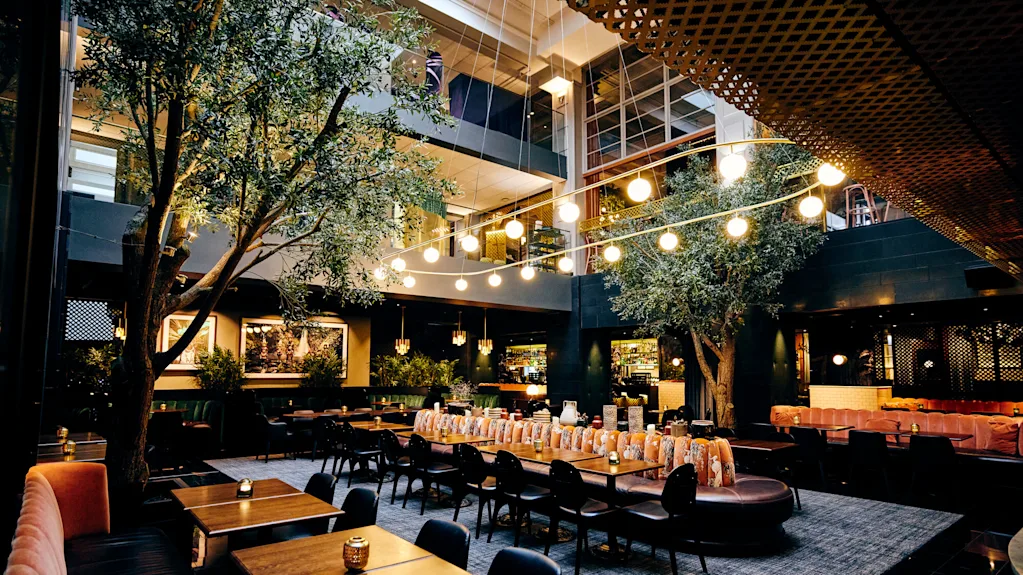 Dining tables sit indoors beneath glowing hanging lights, surrounded by greenery and modern furnishings, creating a warm, inviting restaurant atmosphere.