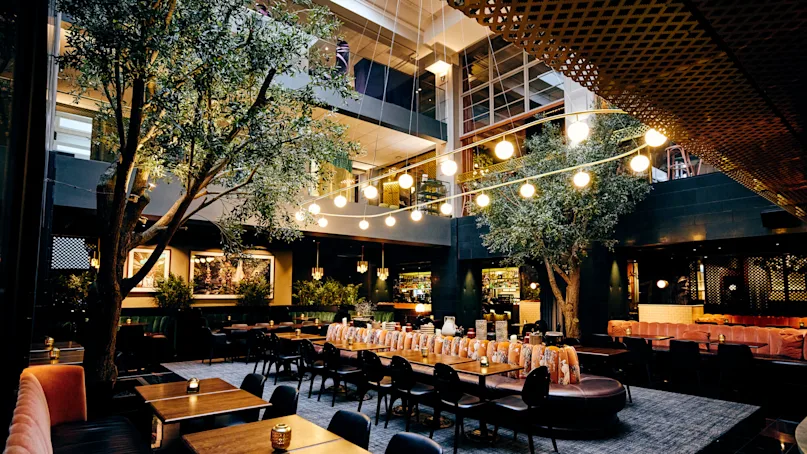 Dining tables sit indoors beneath glowing hanging lights, surrounded by greenery and modern furnishings, creating a warm, inviting restaurant atmosphere.