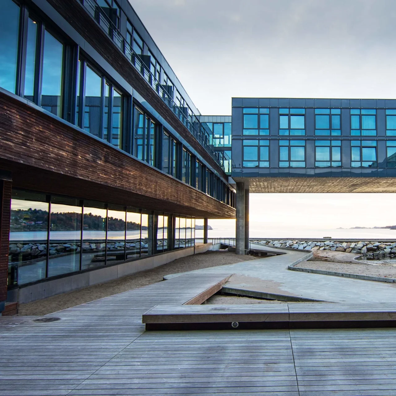 Moderne bygning med en brosektion over en træpromenade ved vandet, tidlig aftenlys, roligt hav i baggrunden.