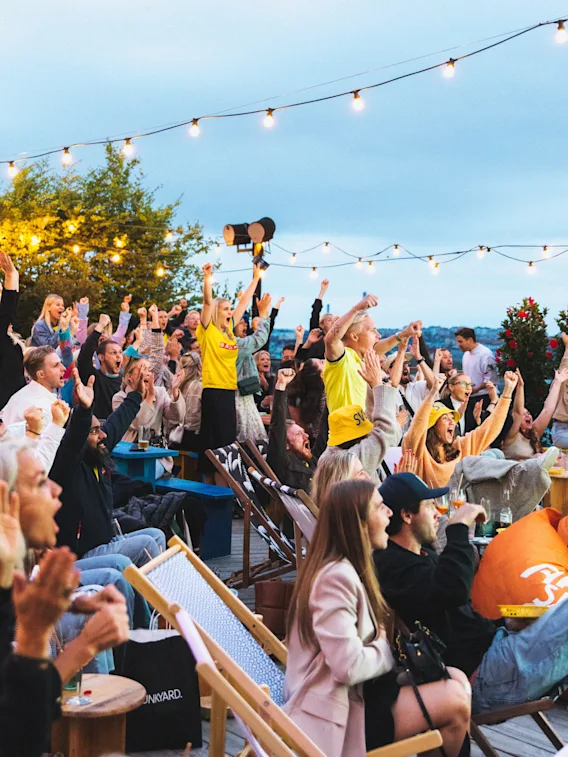 A lively crowd cheers with raised arms at an outdoor event under string lights, Stockholm Under Stjärnorna.