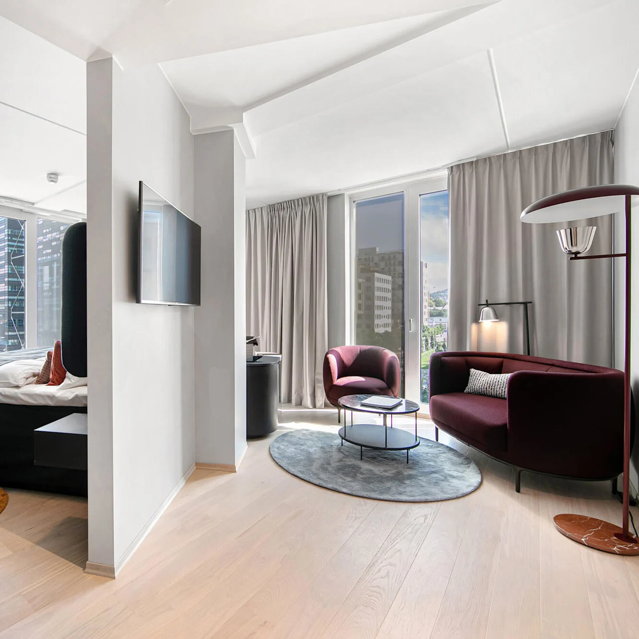 Bedroom with a modern bed, facing a flat-screen TV; adjacent living area features maroon seating, a floor lamp, and round tables. Large windows with city skyscrapers visible outside.